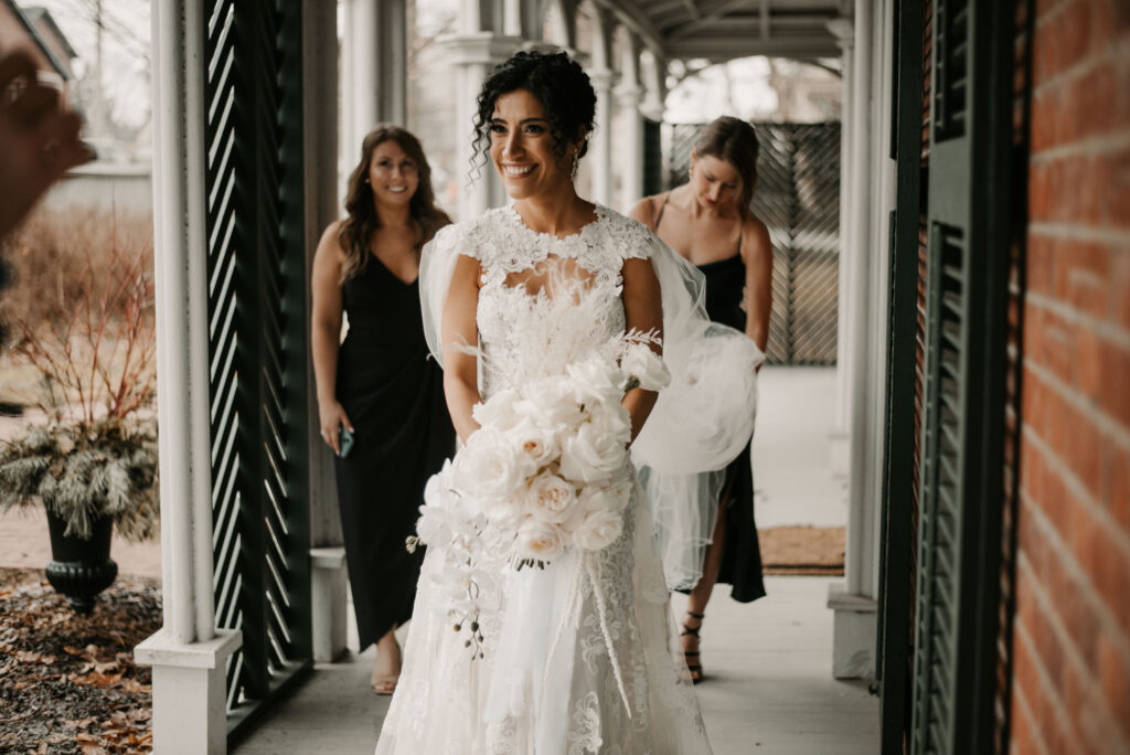 A bride dressed in white, holding a bouquet is smiling to someone off-camera as she walks on the Hillary House verandah, as two bridesmaids dressed in black are holding the dress’s train behind her.